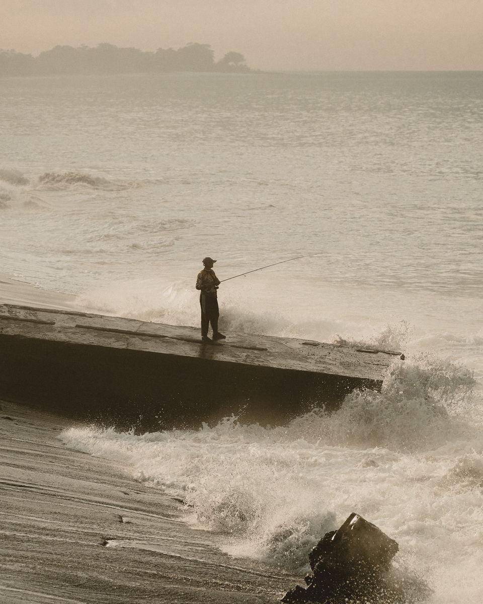 Lone angler casting from a concrete groyne as a wave breaks, sepia morning haze