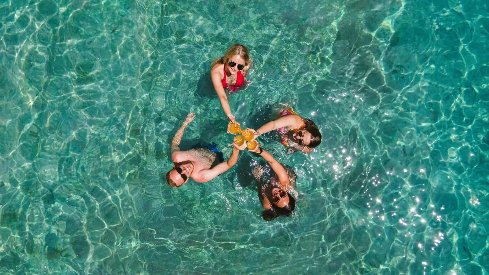 Four guests clinking orange cocktails in a turquoise pool, seen from above